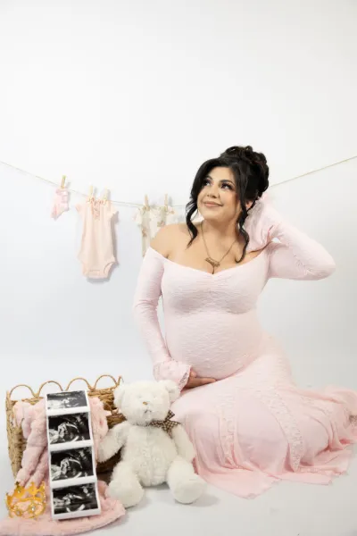 Pregnant woman in pink off-shoulder dress poses with ultrasound photos, teddy bear, and baby clothes on clothesline.