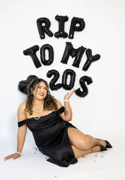 Woman in black dress sitting beside RIP TO MY 20S balloon letters birthday photo.