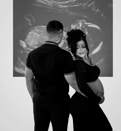 Black and white photo of pregnant couple embracing in front of projected ultrasound image.
