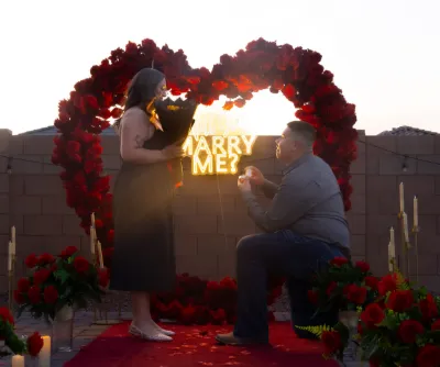 Romantic outdoor marriage proposal with a heart-shaped rose arch and glowing Marry Me sign at sunset.