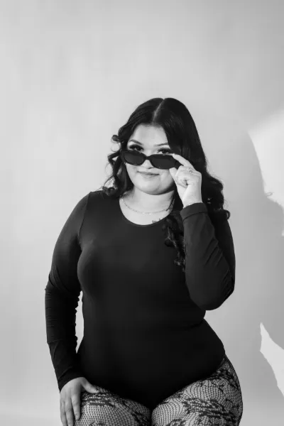 Plus-size woman in black bodysuit holding sunglasses in black and white studio photo.
