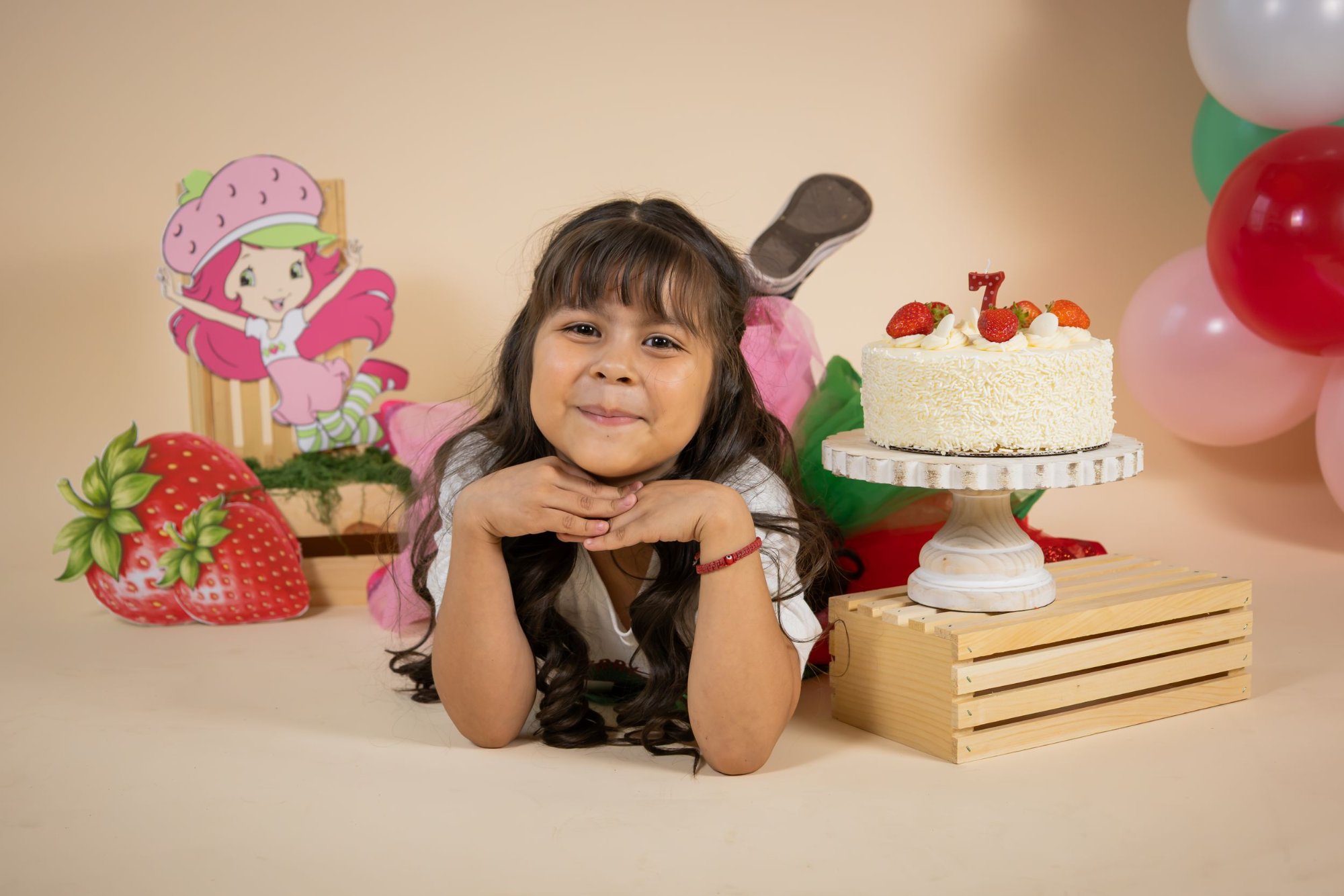 Young girl poses with Strawberry Shortcake themed birthday cake and decorations celebrating her 7th birthday.