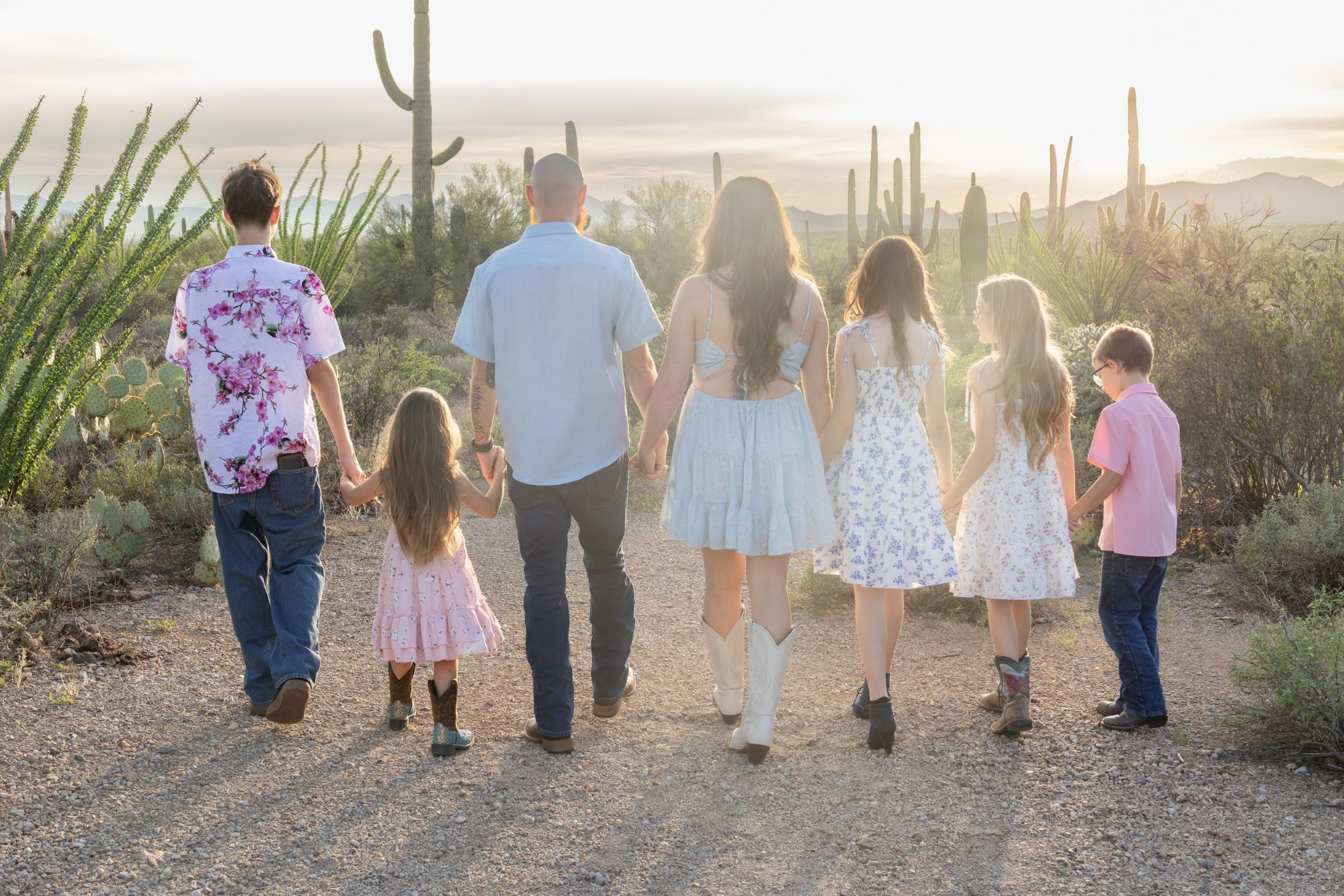 Family of seven holding hands walking on a desert path surrounded by cacti at sunset.