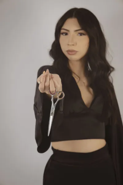 Woman in black crop top holding silver scissors toward camera with long wavy dark hair.