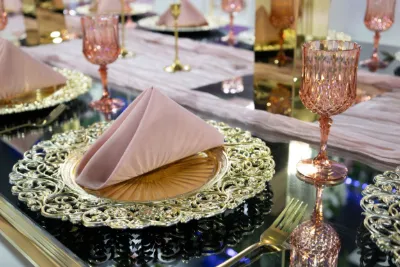 Elegant gold charger plates with pink folded napkins and rose-colored crystal glasses on mirrored table.