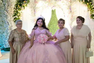 Young girl in a pink quinceañera ball gown poses with three women in front of a floral backdrop.