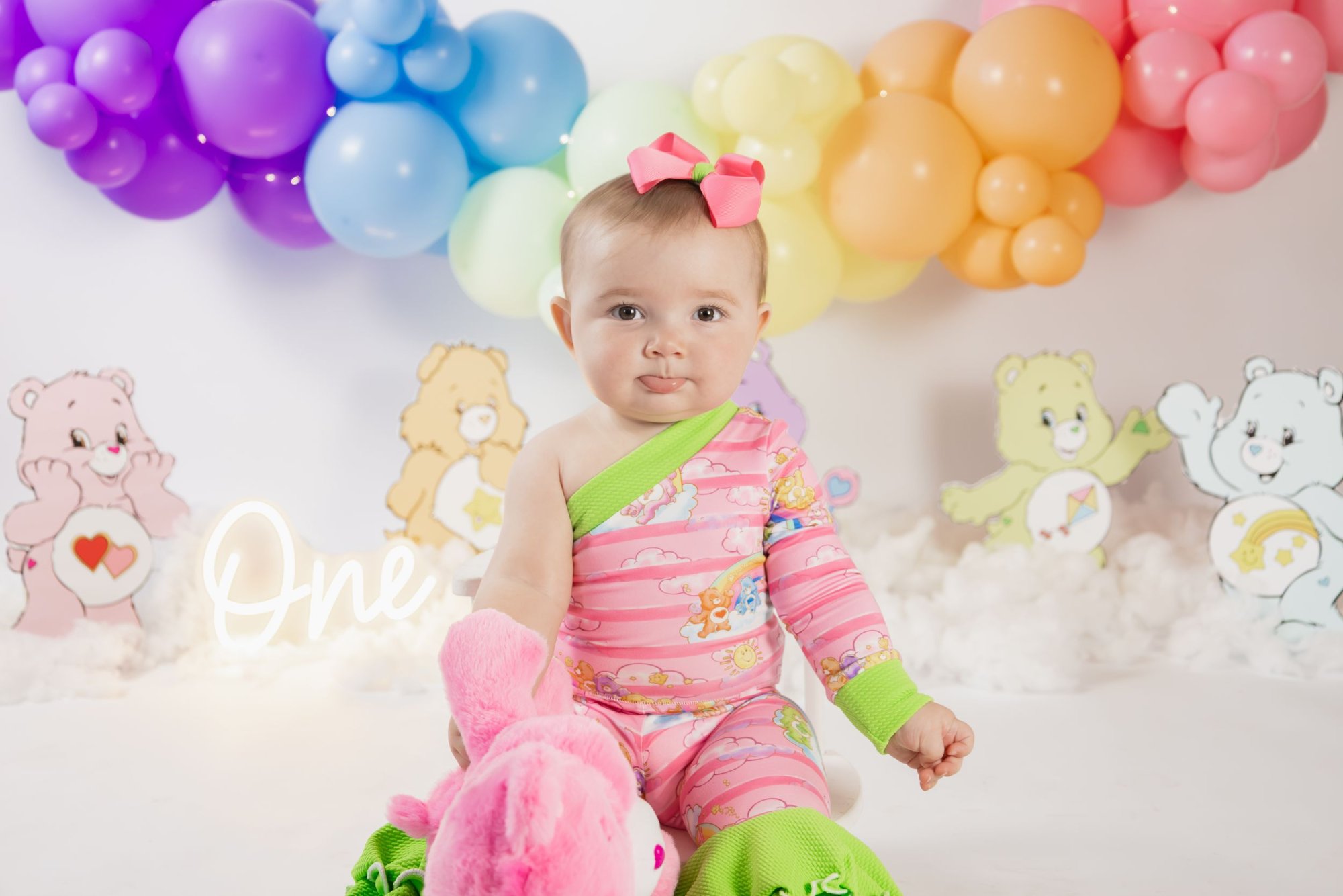 Baby girl in pink Care Bears outfit surrounded by rainbow balloons and Care Bear decorations.