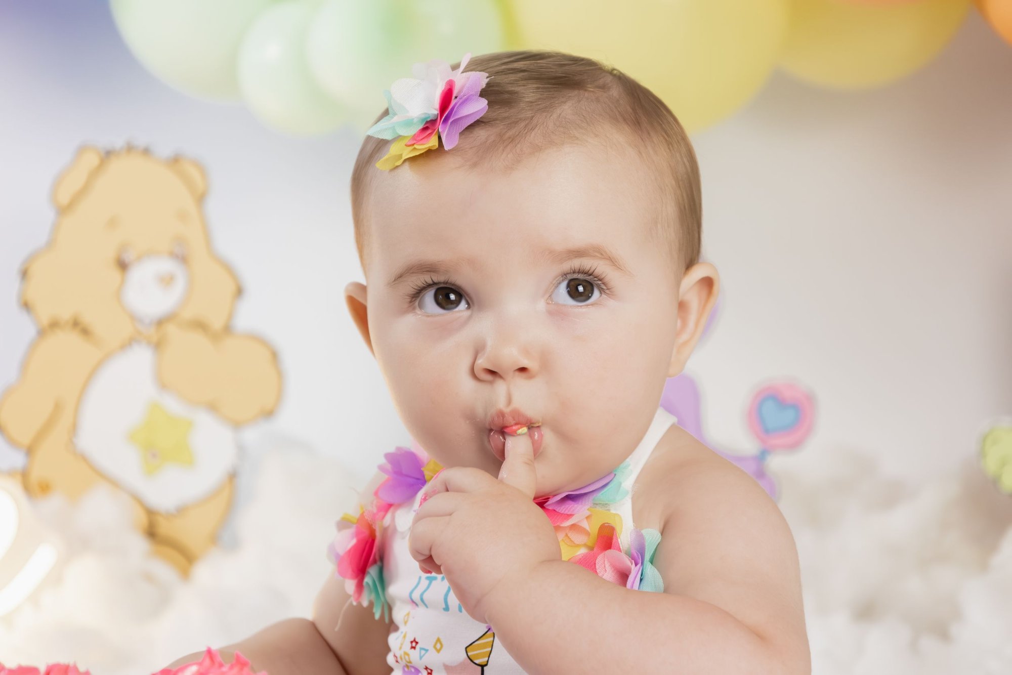 An adorable baby girl with a colorful bow headband at a Care Bears themed first birthday celebration.
