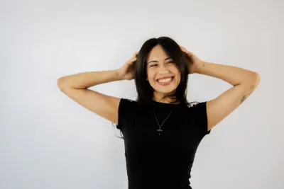 Smiling young woman in black t-shirt with hands in dark hair against white background.