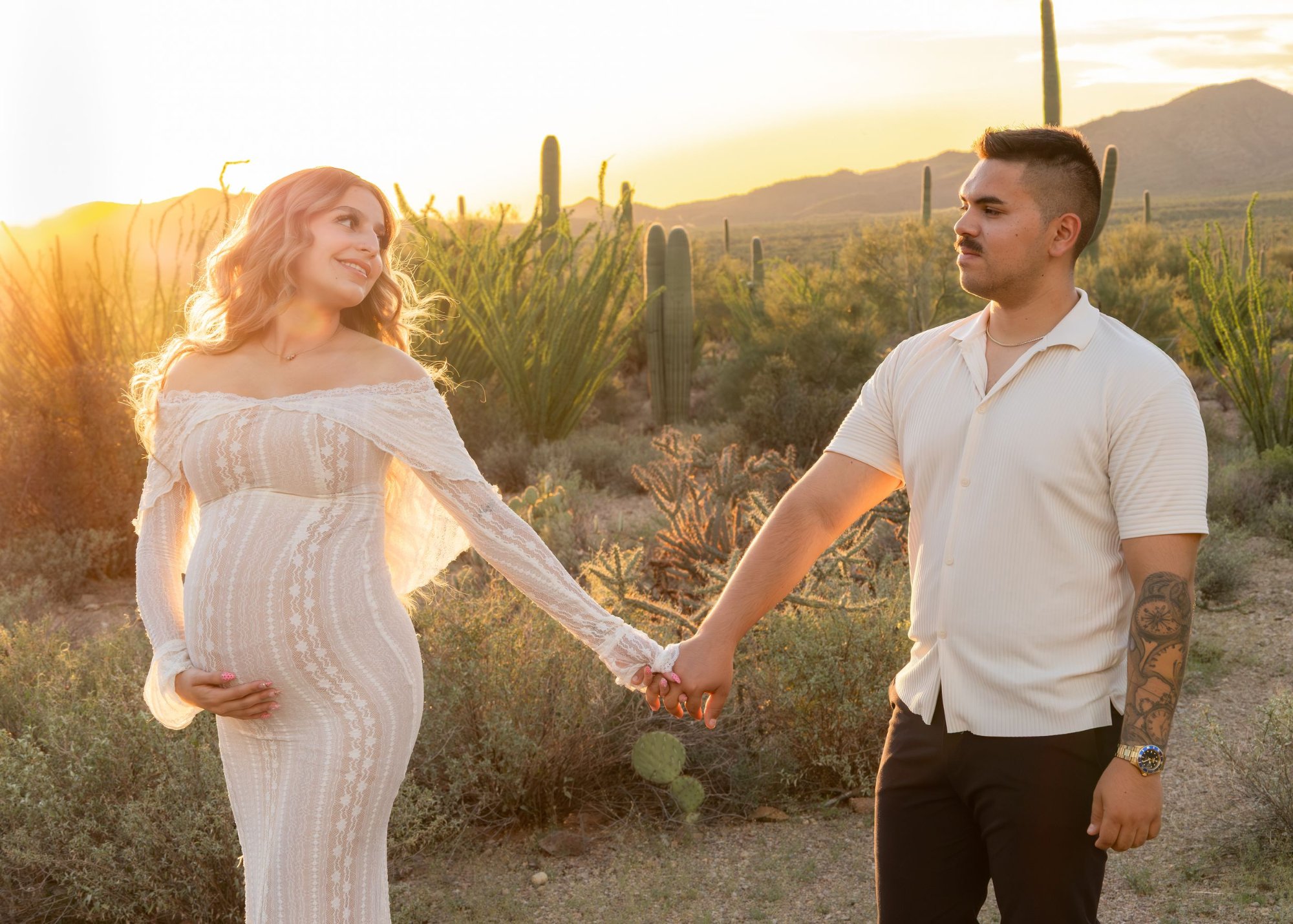 Pregnant couple holds hands at golden sunset in desert landscape with saguaro cacti.