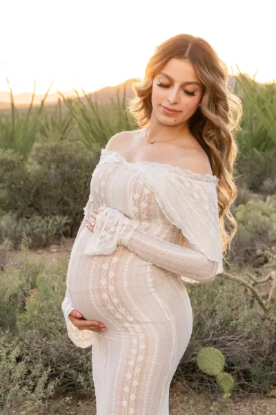Pregnant woman in white off-shoulder lace gown holding baby bump at golden sunset in desert landscape.