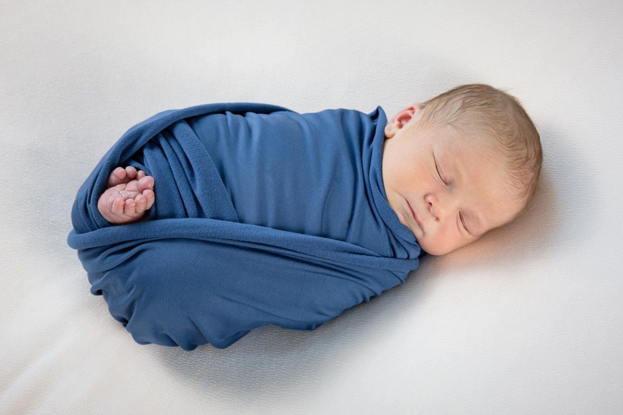 Peaceful newborn baby swaddled in a blue wrap sleeping on a soft white surface.