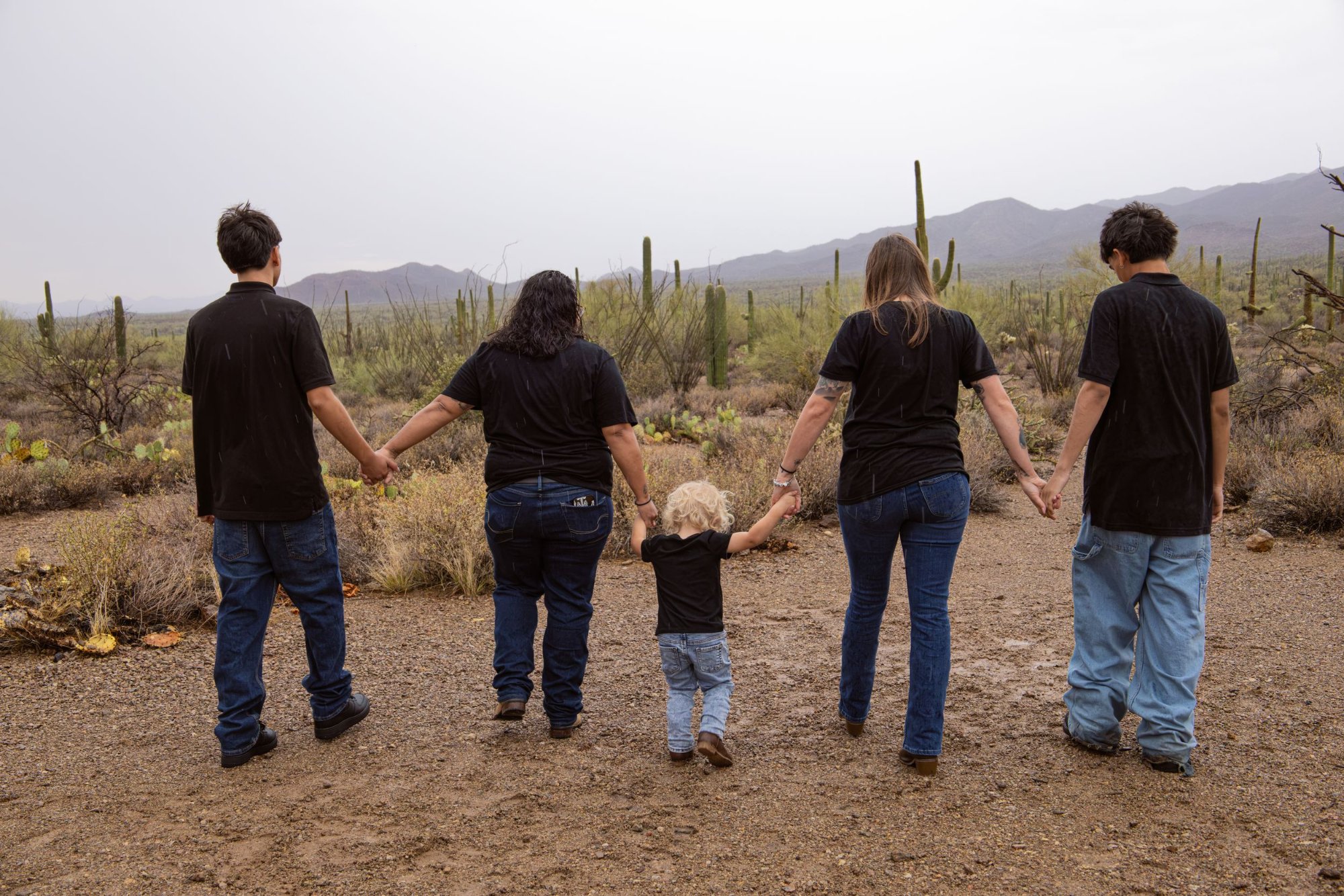 Family of five in black outfits holding hands while walking in a desert landscape with saguaro cacti.