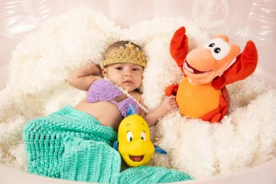 Baby dressed as Little Mermaid Ariel with Sebastian and Flounder plush toys in white setting.