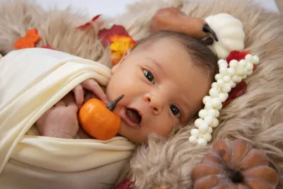 Newborn baby in cream wrap posed on fur with mini pumpkins and fall leaves for autumn photo shoot.