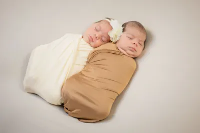 Two sleeping newborn twins swaddled in cream and tan wraps, with one wearing a white flower headband.