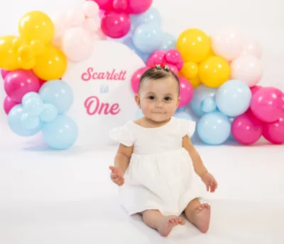 Baby girl in white dress at first birthday celebration with colorful balloon arch backdrop.