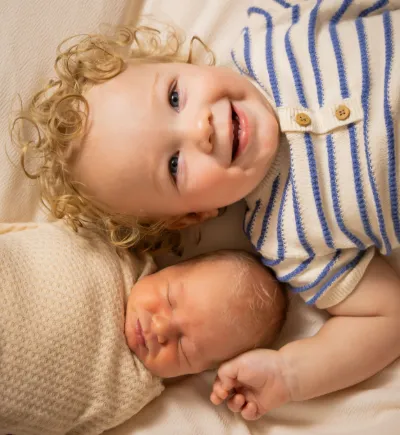 Smiling toddler with curly blonde hair poses with sleeping newborn baby wrapped in cream blanket.