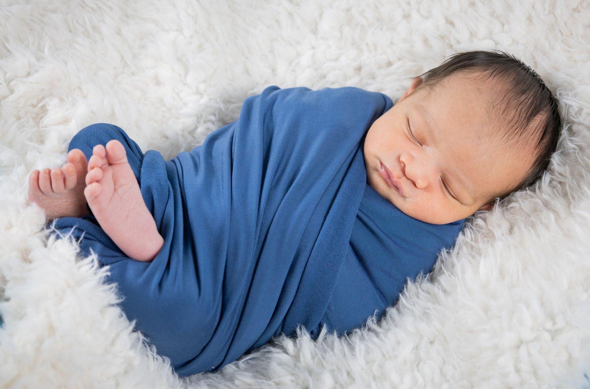 Sleeping newborn baby swaddled in a blue wrap on a white fluffy blanket.