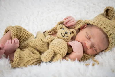 A sleeping newborn in a knit bear outfit cuddles a matching teddy bear on a soft white blanket.