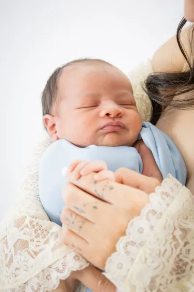 Mother in lace dress tenderly holding sleeping newborn baby wrapped in light blue swaddle.