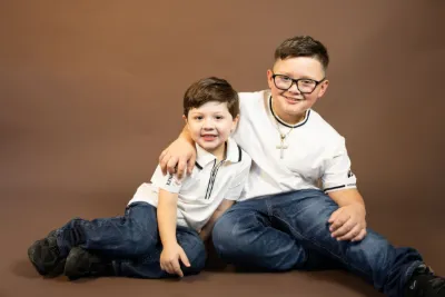 Two young brothers in white shirts and jeans pose together smiling against a warm brown studio background.