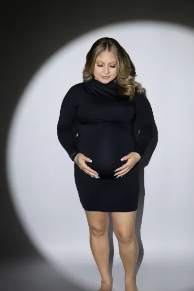 Pregnant woman in a fitted black turtleneck dress cradling her baby bump in a studio spotlight setting.