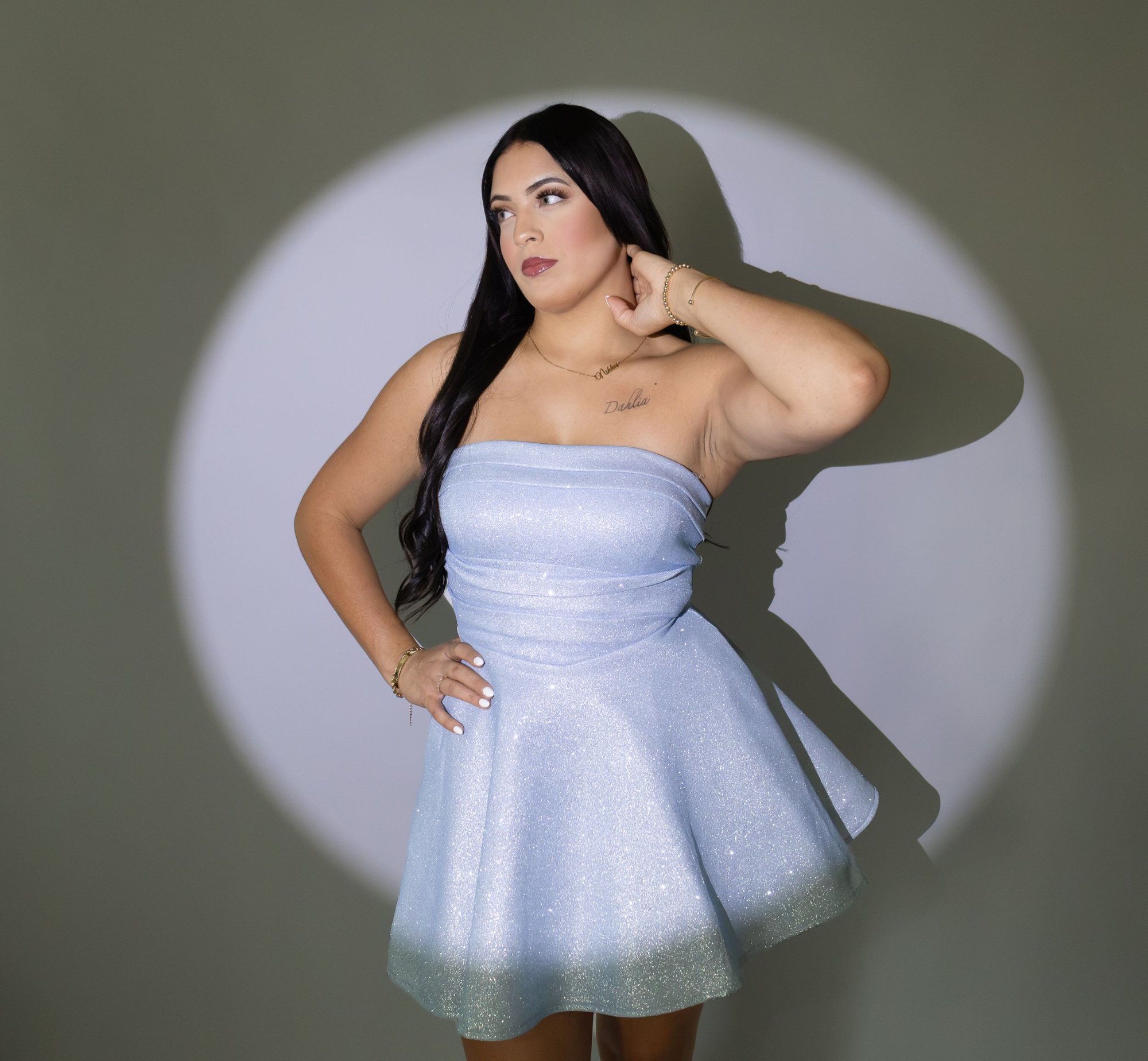 Plus-size woman posing in strapless sparkly light blue mini dress under a spotlight against green background.