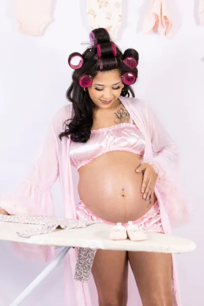 Pregnant woman in pink lingerie and hair rollers cradling her baby bump near an ironing board with baby clothes.