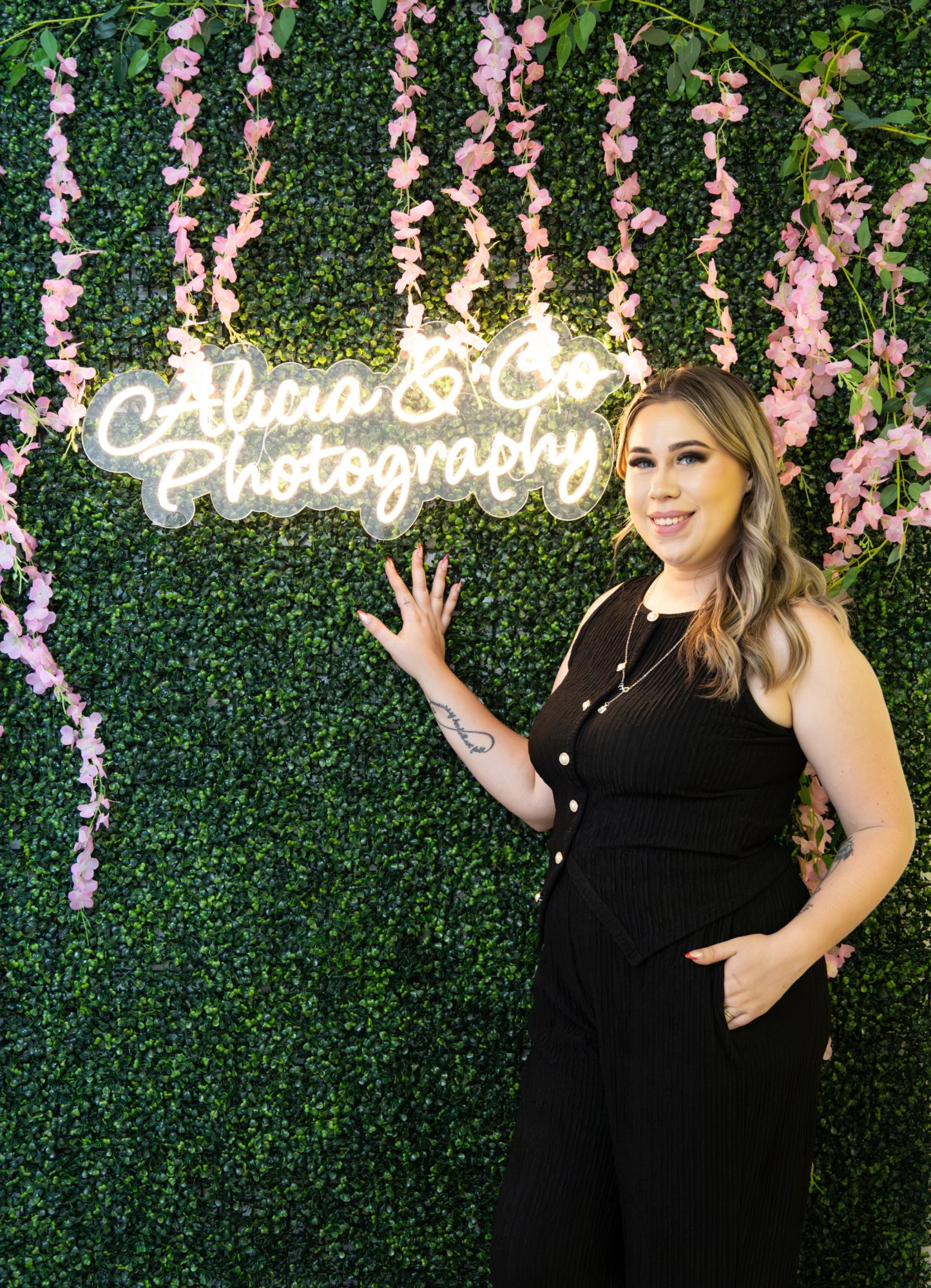 Alicia beside the Alicia & Co Photography neon sign on the green boxwood wall in her studio.