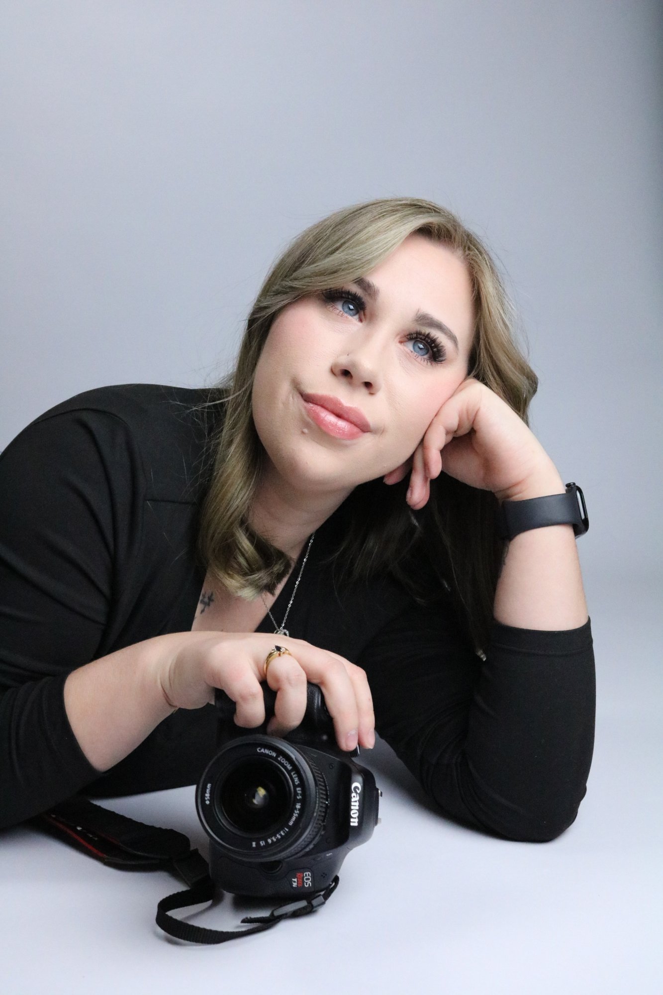 A blonde woman in black clothing posing with a Canon DSLR camera against a gray studio background.