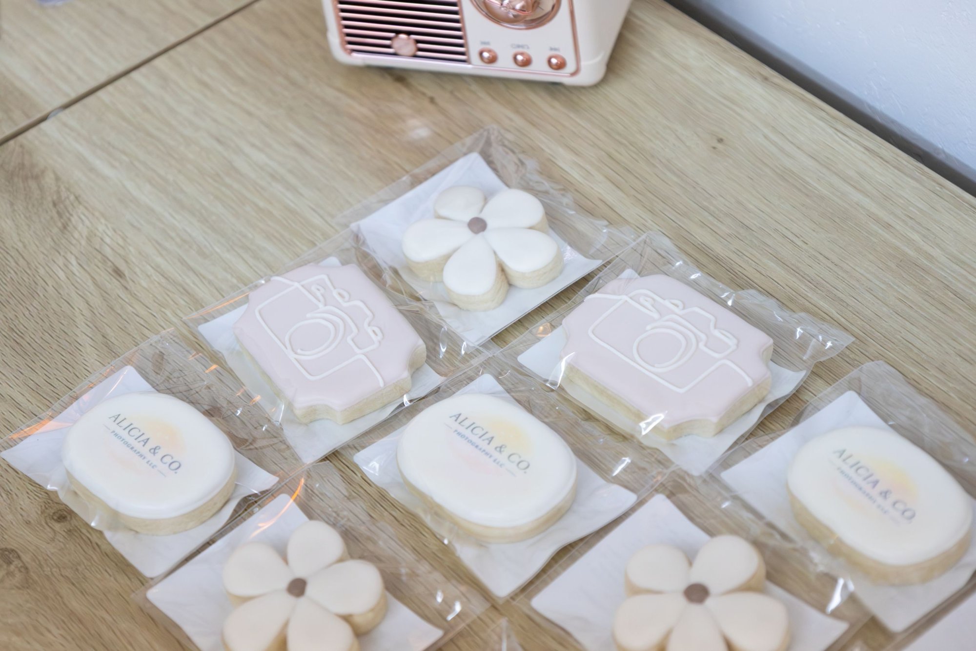 Individually wrapped Alicia & Co. branded cookies with the studio logo, a line-drawn camera and daisy designs, arranged on a wooden table.