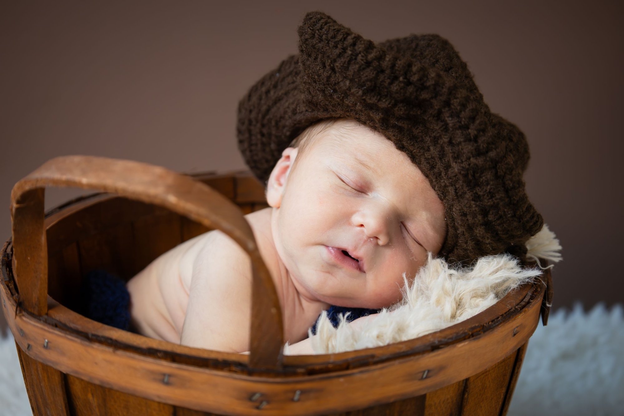 Sleeping newborn baby wearing brown crocheted cowboy hat nestled in rustic wooden basket with white fur.